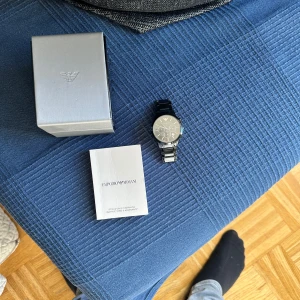 Svart klocka från Emporio Armani - Säljer en snygg svart klocka från Emporio Armani. Klockan har en stilren design med svart urtavla och silverdetaljer. Den kommer med originalförpackning och manual. Perfekt för både vardag och fest!
