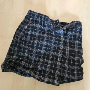 Svart och vit rutig kjol - Säljer en snygg svart och vit rutig kjol med knappar framtill. Kjolen är perfekt för både vardag och fest. Den har en normal passform och är superbekväm att ha på sig. Passar bra till en enkel topp eller en stickad tröja för en mer avslappnad look.