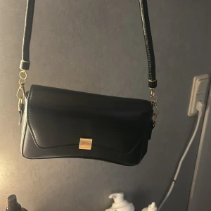 Svart axelväska med gulddetaljer - Säljer en snygg svart axelväska i skinn med gulddetaljer. Väskan har en elegant design med en liten guldfärgad spänne på framsidan. Perfekt för både vardag och fest!