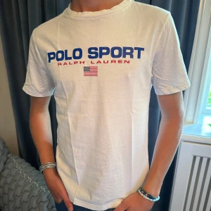 Vit t-shirt från Ralph Lauren - Säljer en snygg vit t-shirt från Ralph Lauren med texten 'Polo Sport' i blått och 'Ralph Lauren' i rött på bröstet. Den har också en liten amerikansk flagga under texten. Perfekt för en casual look! 💙