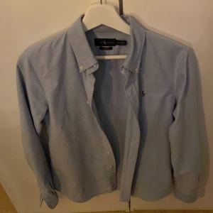 Blå skjorta från Ralph Lauren - Snygg blå skjorta från Ralph Lauren med broderad logotyp på bröstet. Skjortan är knappt använd.  Perfekt för en stilren look. Dåligt ljus på bilden ser mycket bättre ut i verkligheten😄