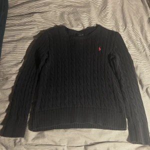 Mörkblå stickad tröja från Ralph Lauren - Säljer en klassisk mörkblå stickad tröja från Ralph Lauren med kabelstickat mönster. Tröjan har långa ärmar och en liten röd logotyp på bröstet. Perfekt för kyligare dagar.