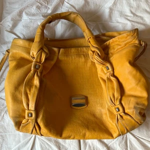 Gul väska från Francesco Biasia - Snygg gul väska från Francesco Biasia med knutna handtag och ett mönstrat innerfoder. Väskan har en dekorativ metallplatta med logotyp på framsidan och är tillverkad i ett syntetmaterial med krokodilmönster. Den har tappat lite gul färg. Pris kan diskuteras