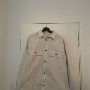 Beige overshirt från H&M - Snygg vit/beige overshirt från H&M med relaxed fit. Den har långa ärmar och knappar framtill samt två stora bröstfickor med knappar. Perfekt för lager-på-lager-stil.