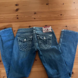 Blå jeans från True Religion - Snygga blå jeans från True Religion med kontrasterande vita sömmar och klassiska bakfickor med lock och knappar. Jeansen har en låg midja och en straight passform, perfekt för en avslappnad stil.
