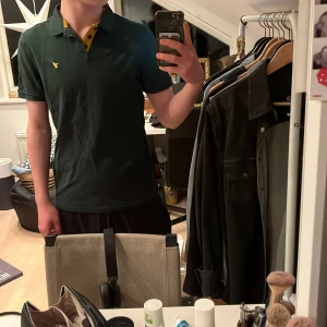 Grön pikétröja från Pier One - Snygg grön pikétröja från Pier One med gul detalj vid kragen och en broderad logga på bröstet. Perfekt för en avslappnad stil.