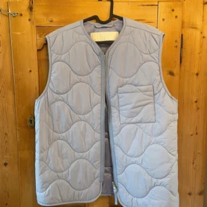 Arket väst - Säljer en ljusblå quiltad väst från Arket med dragkedja framtill och en praktisk bröstficka. Västen har ett vågigt mönster och är perfekt för lager-på-lager-stil. Den är ärmlös och har en rundad halslinje. Skicket är verkligen precis som ny, har bara använt den 2 gånger, säljer pga att den är för stor för mig. Tveka inte med att föreslå pris!
