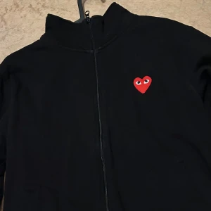 Cdg zip - Snygg svart hoodie med dragkedja från Comme des Garçons. Den har ett rött hjärta med ögon på bröstet och flera hjärtan på ryggen tillsammans med texten 'PLAY'. Perfekt för en stilren och trendig look.
