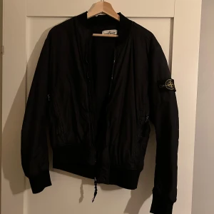 Svart bomberjacka från Stone Island - Snygg svart bomberjacka från Stone Island med dragkedja och ribbade muddar. Jackan har en ikonisk patch på ärmen och är perfekt för en stilren look.