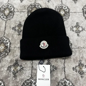 Moncler mössa - Snygg svart stickad mössa från Moncler med ribbad design och klassisk logga framtill. Perfekt för kyliga dagar och en stilren look.