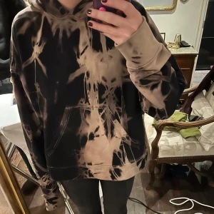 Svart och beige hoodie med batikmönster - Cool hoodie i svart och beige med ett unikt batikmönster. Den har en stor ficka framtill och en bekväm huva. Det är bra kvalite och den har en tyngd till sig. Jag skulle säga att det är en strl L men jag själv bär S/M så den blir lite oversized. 