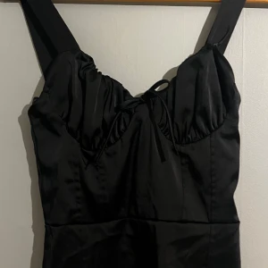 Svart mini klänning med knytband - Elegant svart klänning med breda axelband och knytband framtill. Den har en smickrande passform och smockdetaljer på baksidan för extra komfort. Perfekt för en stilren look.