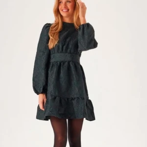 Mörkgrön klänning med volangdetaljer - Nypris 900kr! Är i nyskick och prislapp är kvar! Elegant mörkgrön klänning med långa ärmar och volangdetaljer. Klänningen har en struktur med blommigt mönster och en markerad midja. Perfekt för höst och vinter!