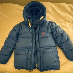 Blå dunjacka från Fjällräven - Säljer en blå dunjacka från Fjällräven med huva och praktiska fickor. Jackan har en dragkedja och knappar framtill samt en broderad logotyp på bröstet. Perfekt för kalla dagar! Jätte mysig och har fin färg.