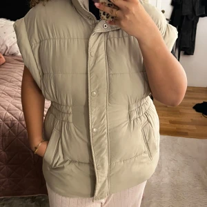 Beige dunväst från Gina Tricot - Säljer en snygg beige dunväst från Gina Tricot i storlek M. Västen har en dragkedja och knappar framtill samt elastisk midja för en bekväm passform. Perfekt för kyligare dagar!