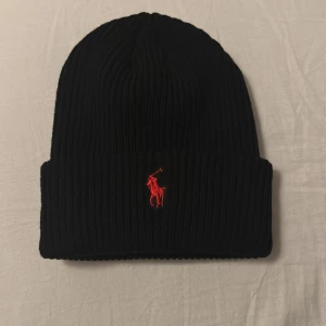 Svart Ralph Lauren mössa - Snygg svart mössa från Polo Ralph Lauren med röd broderad logga framtill. Perfekt för att hålla värmen under kyliga dagar nu i vintern. Den är i nyskick och har knappt används. Pris t kan diskuteras!