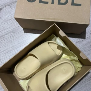 YEEZY Bone (2022 Restock) tofflor - Säljer ett par bekväma beige sandaler från YEEZY adidas  Perfekta för avslappnade dagar. De har en enkel design med öppen tå och är tillverkade i syntetmaterial.