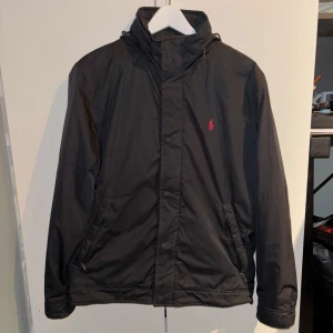 Svart jacka från Ralph Lauren - Snygg svart jacka från Ralph Lauren med röd broderad logga på bröstet. Jackan har en hög krage och dragkedja framtill. Perfekt för kyligare dagar. Priset kan diskuteras!