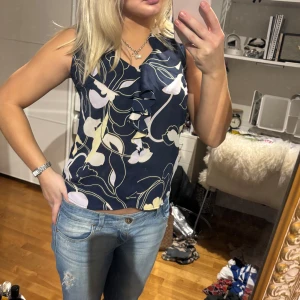 Blommig ärmlös topp - Snygg ärmlös topp med blommigt mönster i blått, vitt och gult. Toppen har en dekorativ volangdetalj framtill och en bekväm passform. 