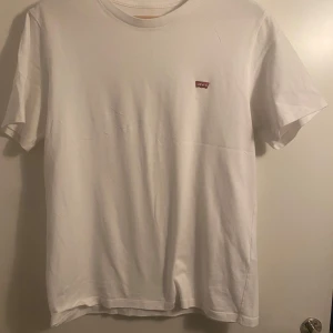 Vit t-shirt från Levi's - Säljer en klassisk vit t-shirt från Levi's med deras ikoniska röda logga på bröstet. T-shirten är kortärmad och tillverkad i mjuk bomull, perfekt för en avslappnad stil.