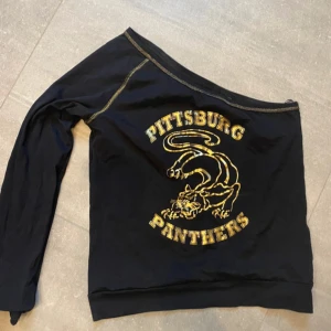 Svart offshoulder tröja med Pittsburg Panthers-tryck - Snygg svart offshoulder tröja med gult Pittsburg Panthers-tryck. Perfekt för en avslappnad stil med en unik touch. Tröjan har en lång ärm och en cool design som sticker ut. Strl L men sitter även snyggt på en ca M. Använd gärna köp nu, bundel eller prisförslag 💫