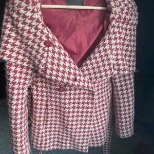 Rutig kappa med bälte - Snygg rutig kappa från H&M i rött och vitt med ett klassiskt hundtandsmönster. Jackan har långa ärmar och ett bälte i midjan för en figursydd look. Perfekt för höst och vinter med sin stilrena design och stora krage.