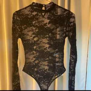 Svart spetsbody - Elegant svart spetsbody med långärmad design. Perfekt för en stilfull look med sin genomskinliga spets och höga hals. Passar utmärkt till både fest och vardag.