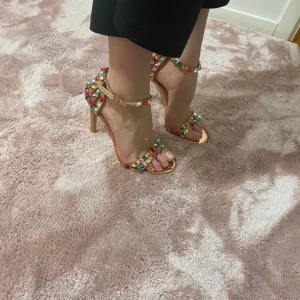 Färgglada klackskor med nitar - Snygga klackskor i guld med färgglada nitar i rött, grönt och blått. Skorna har en öppen tå och justerbar rem runt ankeln. Perfekta för att ge en färgklick till din outfit.