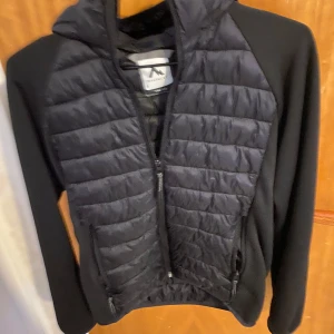 Svart jacka från McKinley - Säljer en svart jacka från McKinley med quiltad design och dragkedja. Jackan har en huva och långa ärmar, perfekt för kyligare dagar. Den är både stilren och funktionell med praktiska fickor framtill.