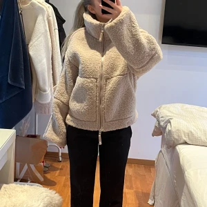 Beige teddyjacka från H&M - Mysig beige teddyjacka från H&M med dragkedja och stora fickor framtill. Perfekt för kyliga dagar med sin fluffiga och varma design. Jackan har en hög krage och långa ärmar för extra komfort.