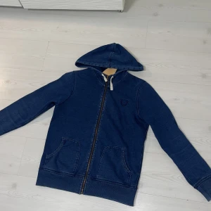 Blå huvtröja från True Religion - Säljer en blå huvtröja från True Religion med dragkedja och två fickor framtill. Tröjan har en justerbar huva och är tillverkad i ett mjukt material för extra komfort. Perfekt för en avslappnad stil. Köpt för ungefär 1400kr och säljer den för endast 850kr, kom gärna med frågor och prisförslag.