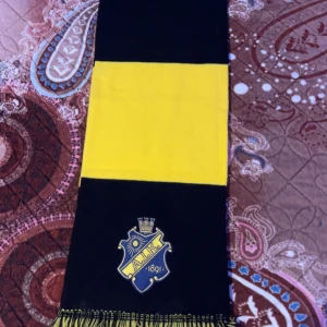 Svart och gul halsduk med AIK-logga - Snygg halsduk i svart och gult med AIK-logga. Perfekt för att visa ditt stöd på matchen. Den har fransar i ändarna och är gjord i ett mjukt material.