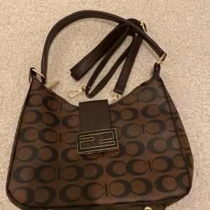 Brun mönstrad axelväska - Snygg brun axelväska med ett stilrent mönster och gulddetaljer. Väskan har en praktisk dragkedja och en justerbar axelrem. Perfekt för att bära dina dagliga nödvändigheter med stil.