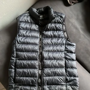 Mörk blå  dunväst från Uniqlo - Säljer en stilren mörk blå dunväst från Uniqlo. Västen är ultralätt och perfekt för lager-på-lager under kyliga dagar. Den har en dragkedja framtill och är quiltad för extra värme. Passar perfekt för både höst och vår.