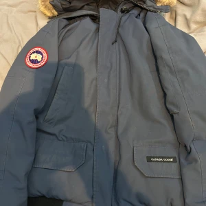 Blå parkajacka från Canada Goose - Säljer en blå parkajacka från Canada Goose med pälsdetaljer på huvan. Jackan har en broderad logotyp på ärmen och flera praktiska fickor framtill. Perfekt för kalla vinterdagar.