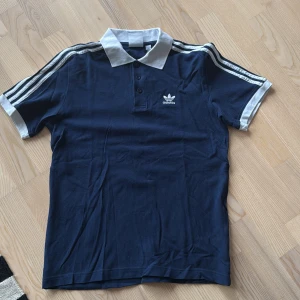 Mörkblå pikétröja från Adidas - Säljer en snygg mörkblå pikétröja från Adidas med vita detaljer och klassiska ränder på ärmarna. Tröjan har en vit krage och knappar framtill. Perfekt för en sportig look!