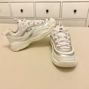 Vita sneakers från Fila - Snygga vita sneakers från Fila med silverdetaljer och röd-vit snörning. Perfekta för en sportig look med en chunky sula och bekväm passform.