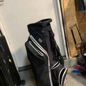 Svart golfbag från Wilson - Säljer en stilren svart golfbag från Wilson med vita detaljer. Bagen har flera fack med dragkedjor för praktisk förvaring av golfutrustning. Perfekt för golfentusiasten som vill ha en pålitlig och snygg bag på banan.