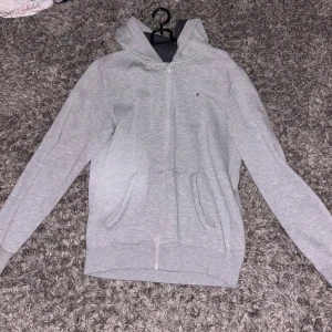 Grå hoodie från Tommy Hilfiger - Säljer en stilren grå hoodie från Tommy Hilfiger med dragkedja och fickor framtill. Den har en liten logga på bröstet och en bekväm huva. Lite nopprig men inga tydliga täcken på användning.