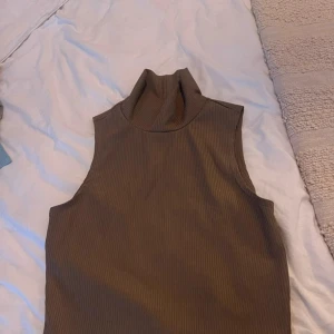 Brun ribbad polotopp från Monki - Säljer en stilren brun ribbad polotopp från Monki. Toppen är ärmlös och har en hög krage, perfekt för lager-på-lager eller som den är. Passar utmärkt till både vardag och fest.