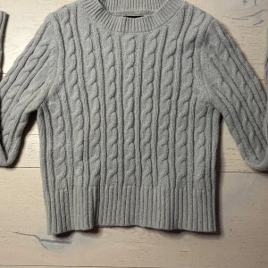 Grå stickad tröja - Säljer en mysig grå stickad tröja med kabelstickat mönster. Tröjan har långa ärmar och ribbstickade muddar vid ärmslut och nederkant. Perfekt för kyliga dagar!