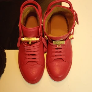Röda sneakers med gulddetaljer - Snygga röda sneakers med snörning och gulddetaljer. Skorna har en unik design med låsdetaljer och är tillverkade i skinn. Perfekta för att ge en färgklick till din outfit.