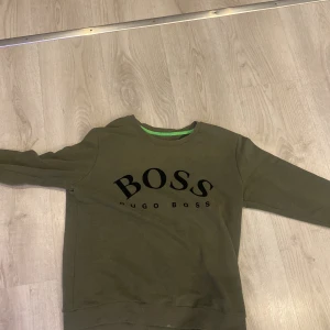 Olivgrön tröja från Hugo Boss - Säljer en snygg olivgrön tröja från Hugo Boss i storlek S. Tröjan har en klassisk design med långärmade ärmar och en rund halsringning. Perfekt för en avslappnad stil eller att klä upp med accessoarer. Boss-loggan pryder framsidan för en stilren look.