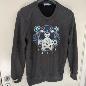 Grå sweatshirt från Kenzo - Säljer en snygg grå sweatshirt från Kenzo med deras ikoniska tigerbroderi i blått och vitt på framsidan. Tröjan har långa ärmar och en rund halsringning. Perfekt för en casual look eller att mysa i hemma. 🐯