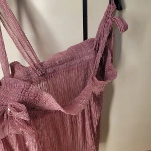 Glittrig rosa top med rosett - Säljer en superfin glittrig rosa top med en söt rosett framtill. Den har en härlig glans och är perfekt för festliga tillfällen. Axelbandslös design med en dekorativ sten i mitten av rosetten. Perfekt för att sticka ut på festen! 💖