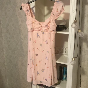 Blommig klänning från Gina Tricot - Söt blommig klänning från Gina Tricot i ljusrosa med små blommor i olika färger. Klänningen har en smickrande passform med volangdetaljer vid axlarna och en kort längd, perfekt för sommarens fester. Materialet känns lätt och luftigt. Säljer för 200