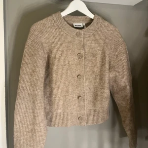 Beige stickad kofta från Weekday - Säljer en superfin beige stickad kofta från Weekday i storlek S. Den har en klassisk design med knappar framtill och ribbade detaljer vid ärmslut och nederkant. Perfekt för höst och vinter, både mysig och stilren. Passar till alla tillfällen! 🍂