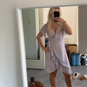 Blommig omlottklänning - Säljer en superfin blommig omlottklänning i ljusa färger. Den har korta ärmar och ett knytband i midjan för en perfekt passform. Perfekt för sommardagar eller en avslappnad vårutflykt. Klänningen är i mycket bra skick och redo för nya äventyr!