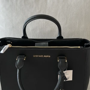 Svart handväska från Michael Kors - Säljer en helt ny elegant svart handväska från Michael Kors i mycket bra skick. Väskan har en stilren design med guldfärgade detaljer och en praktisk dragkedja upptill. Perfekt för både vardag och fest!