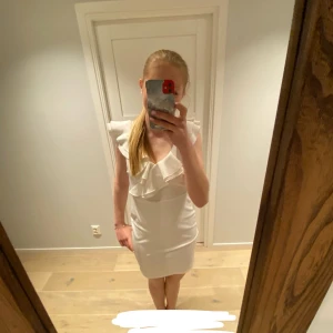Vit klänning från NLY One - Säljer en superfin vit klänning från NLY One i storlek M. Den har en elegant volangdetalj vid halsen och är ärmlös, vilket gör den perfekt för sommarens alla fester. Klänningen har en tight passform som framhäver figuren på ett snyggt sätt. Perfekt för både student och midsommar!